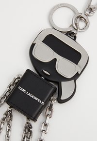 Crni kožni privjesak za ključeve s stiliziranim licem, metalnim sunčanim naočalama, srebrnim dijelovima i lančićem. Ime branda "KARL LAGERFELD" utisnuto.