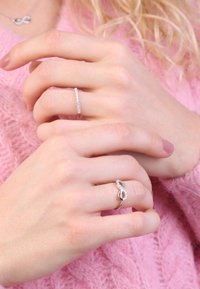 Zwei Hände mit Ringen: ein dünner silberner Ring mit kleinen klaren Steinen und ein silberner Unendlichkeit-Ring mit einem klaren Stein-Akzent. Hellrosa Hintergrund.