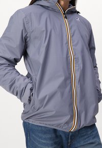 Veste légère grise avec capuche, dotée d'une fermeture éclair frontale avec des rayures d'accent rouges, jaunes et noires. Comprend des poignets élastiques et des poches latérales.