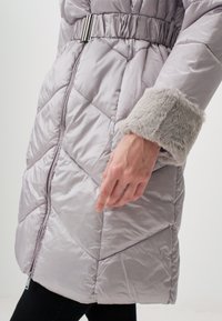 Cappotto imbottito grigio chiaro con cuciture a zigzag, dotato di vita aderente e polsini in ecopelliccia. Il tessuto ha una finitura lucida e una cerniera visibile.