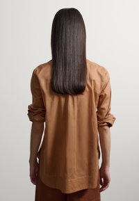 Persona con cabello oscuro, liso y largo, que lleva una camisa beige de manga larga y está de pie de espaldas frente a un fondo claro y liso.
