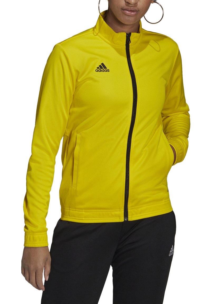 Žuta jakna na zip od glatkog materijala, s visokim ovratnikom i crnim Adidas logotipom na prsima, u kombinaciji s crnim hlačama.