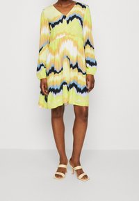 Vestido de manga corta y escote en V, de color amarillo con un patrón de zigzag multicolor. Diseño fluido con longitud por encima de la rodilla, combinado con sandalias de tacón beige.