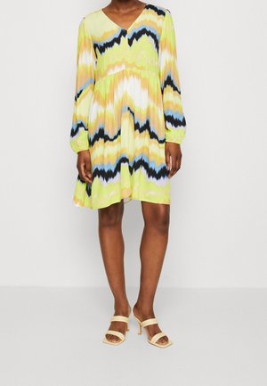 Robe à manches courtes et décolleté en V de couleur jaune avec un motif zigzag multicolore. Design fluide avec une longueur au-dessus du genou, accompagnée de sandales à talons beiges.