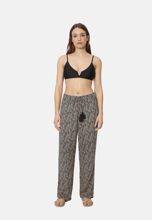 Mujer con bralette negro, pantalones holgados de cintura alta con estampado y lazo de borlas, y sandalias beige, de pie contra un fondo blanco.