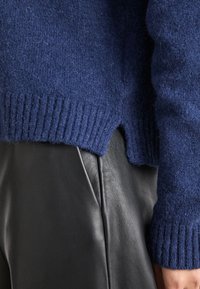 Pull en tricot bleu marine avec un ourlet côtelé, présentant une fente sur le côté. Porté par-dessus un pantalon en cuir noir, mettant en valeur le contraste de textures.