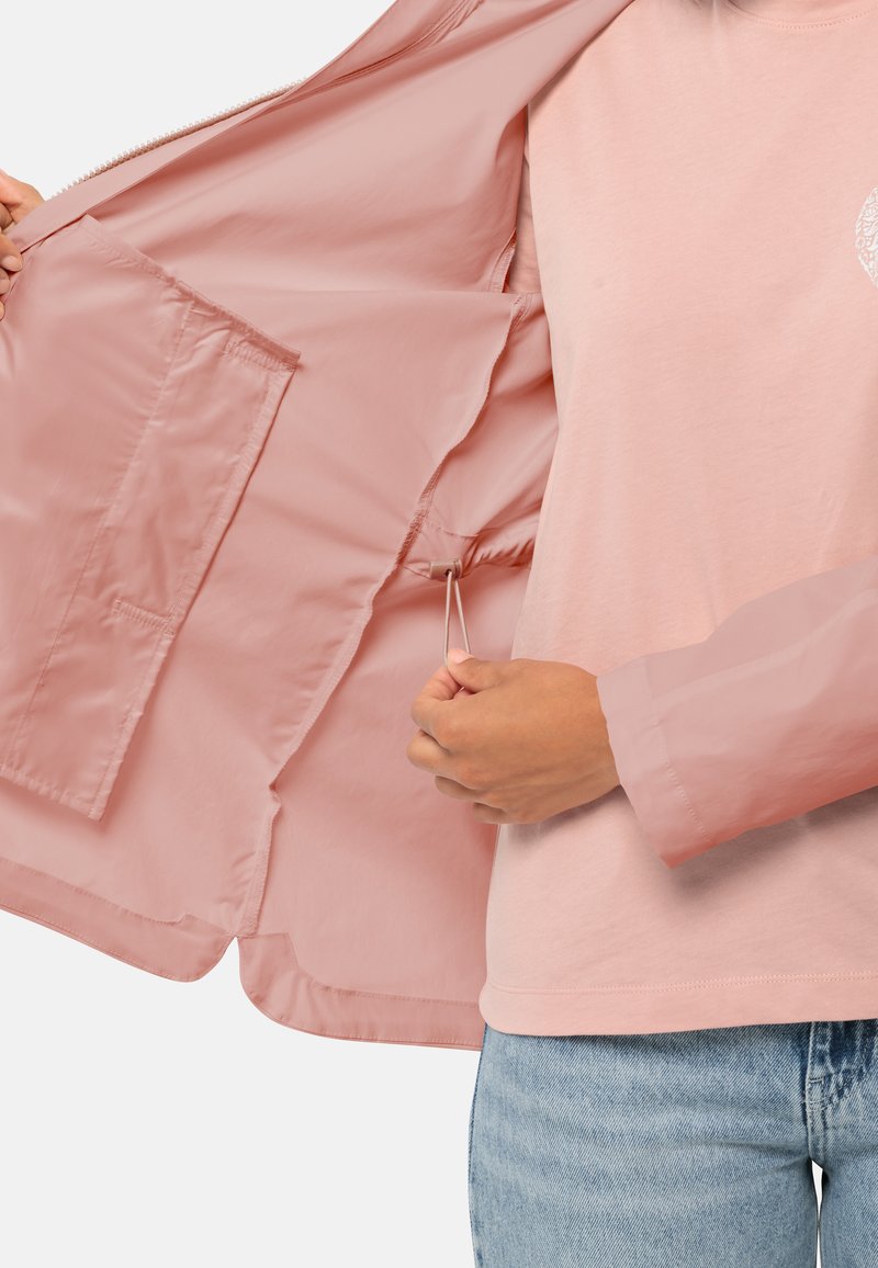 Helle rosa Jacke mit glatter, glänzender Oberfläche und Reißverschluss. Innen hat sie eine Tasche und einen verstellbaren Kordelzug. Sie wird über einem hellen Hemd getragen.