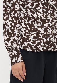 Brun og hvit mønstrete bluse med lange ermer, knappelukking og rynkede mansjetter. Laget av et lett stoff med glatt tekstur.