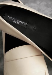 Chaussures en cuir beige à enfiler avec une finition lisse et une large bande sur le dessus. L'intérieur est noir avec une étiquette portant la mention "TABITHA SIMMONS NEXT."