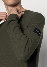 Maglione tricot lungo con maniche a diritta in verde oliva scuro, con texture a costine, caratterizzato da un'etichetta nera Calvin Klein sul polsino e un collo rotondo.