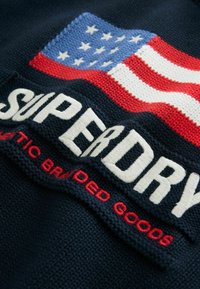Tessuto a maglia navy con un design della bandiera americana nei colori rosso, bianco e blu. "SUPERDRY" è mostrato in modo prominente in ricamo bianco.