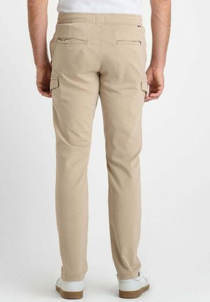 Mann in beigefarbenen Slim-Fit-Hosen mit Rückentaschen und Seitentaschen, weißen Sneakers, stehend vor einem schlichten weißen Hintergrund.