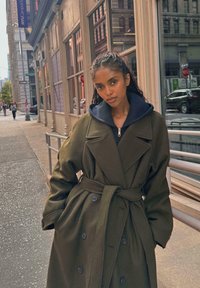 Olivovo zelený dvojradový kabát s veľkými klopami a opaskom. Pod ním sa nachádza námornícky kapucňa a hladká textúra. Mestské pozadie.