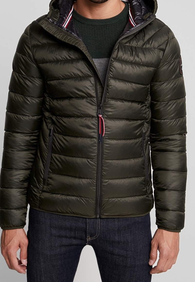Homme portant une doudoune matelassée vert foncé avec capuche, zippée à mi-chemin, par-dessus un pull en maille vert et un jean foncé.