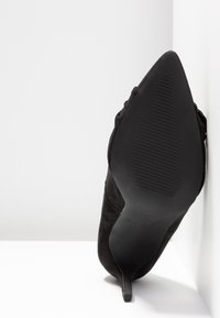 Chaussure à talon aiguille noire en daim à bout pointu, dotée d'un talon fin, d'une semelle en caoutchouc texturé et d'un design minimaliste.