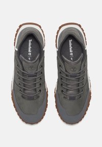 Zapatillas Timberland grises con materiales mixtos, que presentan una superficie texturizada, cordones redondos y una suela de goma marrón. Logo visible en la lengüeta.