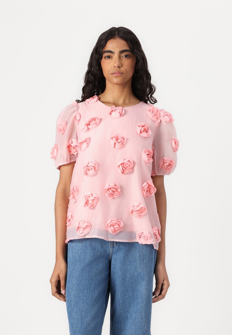 Blusa rosa trasparente decorata con appliqué di rose 3D, maniche a sbuffo e scollatura rotonda. Indossata sopra jeans di denim blu.