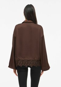 Brun satinbluse med avslappet passform, har klokkeformede ermer og en blonderkant nederst. Blonden har et blomsterpatron og gir en teksturert detalj.