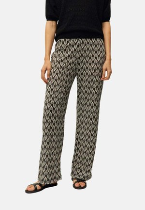 Persona que lleva pantalones anchos con patrón en blanco y negro, camiseta negra de manga corta y sandalias negras con tiras cruzadas.