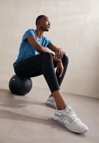 Black leggings and a blue athletic t-shirt on a person sitting on a black medicine ball, wearing light gray shoes with subtle purple accents.