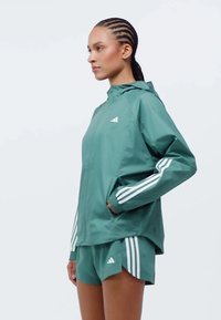 Green waterproof jacket with a hood and white three-stripe accents on the sleeves, paired with matching green shorts. Lightweight material.