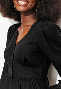 Robe noire avec décolleté en V, détails à boutons et manches bouffantes. Tissu lisse et taille ajustée, s'élargissant en une jupe froncée.