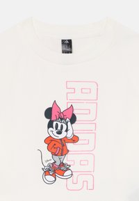 Minnie Mouse oranžā kapučjakā un rozā lentē pozē ar vienu roku uz galvas, blakus rozā tekstam "ADIDAS" uz baltas t-kreklas.