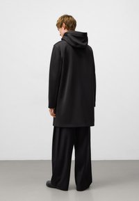 Long manteau noir à capuche en tissu lisse, avec une coupe décontractée et des coutures latérales, associé à un large pantalon noir et des bottes.