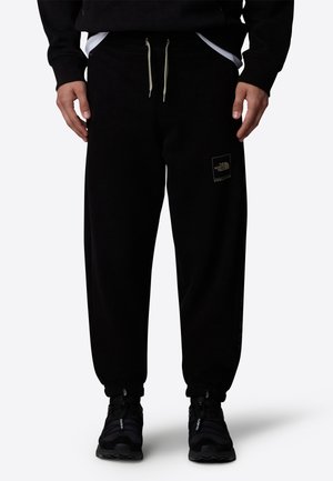 Person trägt schwarze Jogginghose mit The North Face-Logo, schwarze Sneaker und einen schwarzen Pullover, unter dem weiße Saumkanten eines Hemdes sichtbar sind.