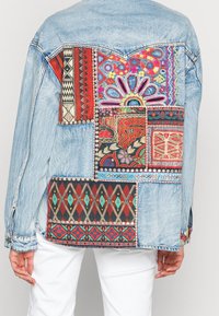 Džínová bunda s barevným patchworkovým zadním dílem, který obsahuje složité geometrické vzory a květinové motivy v červené, modré a krémové barvě, s kontrastním prošíváním.