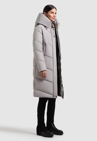 Abrigo acolchado gris con gran capucha, acolchado horizontal, cierres a presión en la parte delantera y bolsillos laterales, llevado sobre un conjunto negro y botas.