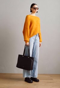 Pull en tricot oversize orange, jeans taille haute bleu clair, sac fourre-tout noir et chaussures noires à semelles épaisses. Le mannequin porte des lunettes de soleil noires.