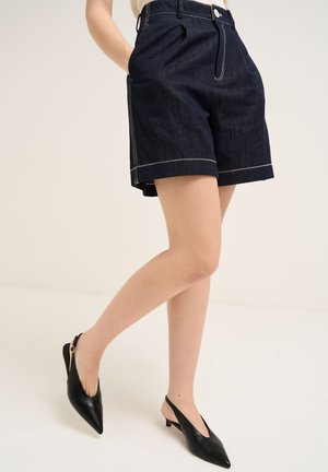 Frau trägt dunkle Jeansshorts mit weißen Nähten und schwarze spitze Slingback-Absätze, steht mit einem leicht gehobenen Bein.
