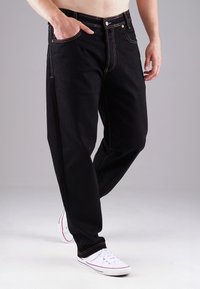 Černé džínové kalhoty s žlutým prošíváním, rovný střih a dvě přední kapsy. Nošeny s bílými teniskami s červenými akcenty.