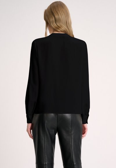 Blusa nera a maniche lunghe realizzata in tessuto morbido, con una vestibilità rilassata e sottili pieghe ai polsini. Abbinata a pantaloni in pelle nera aderenti.
