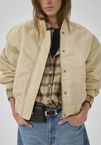 Beige gestepptes Bomberjacke mit Reißverschluss vorne, gerippten Bündchen und lockerer Passform, getragen über einem karierten Flanellhemd, kombiniert mit hochtaillierten Jeans.