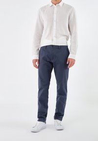 Witte linnen overhemd met knopen, gecombineerd met marineblauwe katoenen broek en witte sneakers. Het shirt heeft een klassieke kraag en lange mouwen.