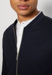 Mornarska bombaška jakna z zadrgo spredaj, izdelana iz teksturirane tkanine. Ima stoječ ovratnik in je prepuščena čez belo srajco z gumbi.