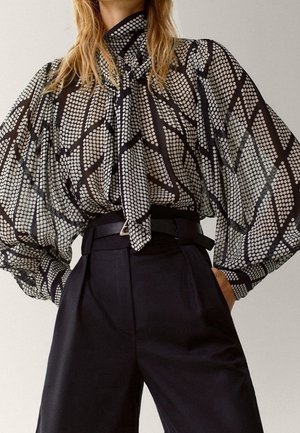 Femme portant un chemisier à motifs noir et blanc avec un nœud papillon au col et un pantalon noir large taille haute.