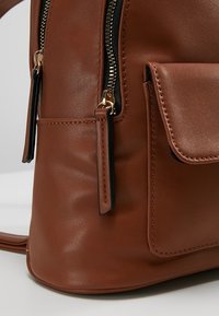 Coin de sac à dos en cuir marron avec tirettes de fermeture éclair dorées et poche avant avec fermeture à rabat cousue.