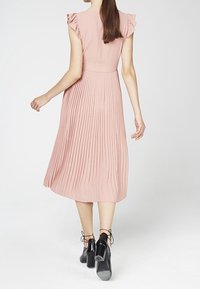 Frau mit langen Haaren, die ein ärmelloses, pinkes, plissiertes Midikleid und schwarze, geschnürte hochhackige Schuhe trägt, die sich von einem weißen Hintergrund wegbewegt.