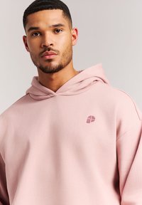 Lichtroze hoodie van zachte stof met een trekkoordkap en een klein logo op de borst. Soepele textuur en ontspannen pasvorm.