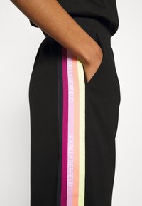 Pantaloni neri con una striscia laterale in tonalità di rosa, viola e giallo con il testo "KARL LAGERFELD". Il tessuto sembra morbido e traspirante.