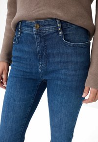Jeans blu scuro con un design aderente, cinque tasche e dettagli in metallo dorato con cuciture visibili. Tessuto testurizzato.