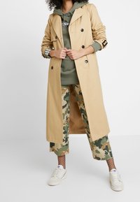 Light beige trench coat with double-breasted buttons, paired with olive green hoodie and camo-patterned pants, white sneakers, and fishnet stockings.