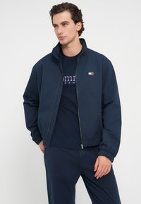 Giacca blu navy con zip, colletto alto, polsini e orlo elastici. Presenta una piccola toppa con logo sul petto. Indossata sopra una maglietta blu navy.