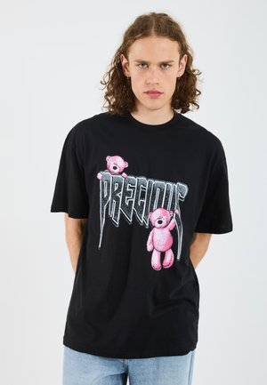 T-shirt en coton noir avec des graphiques de teddy bear rose et le texte "PRECIOUS" en gras, dans une police stylisée. Manches courtes et coupe décontractée.