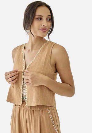 Mujer con chaleco sin mangas color beige con botones y pantalones a juego con bordado blanco, ajustando el chaleco, mirando a su izquierda.