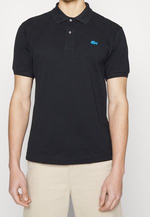 Homme portant un polo à manches courtes noir avec deux boutons et un petit logo crocodile bleu sur le côté gauche de la poitrine.