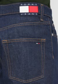 Σκούρα μπλε τζιν υφάσματος denim με πορτοκαλί ραφές, που διαθέτουν ορθογώνιο ένθετο λογότυπο Tommy Jeans με κόκκινες, λευκές και μπλε λεπτομέρειες στη μέση.